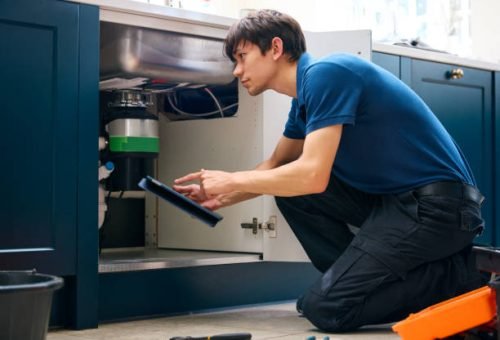 Male Plumber With Digital Tablet Fixing Waste Disposal Unit In Domestic Kitchen Sink